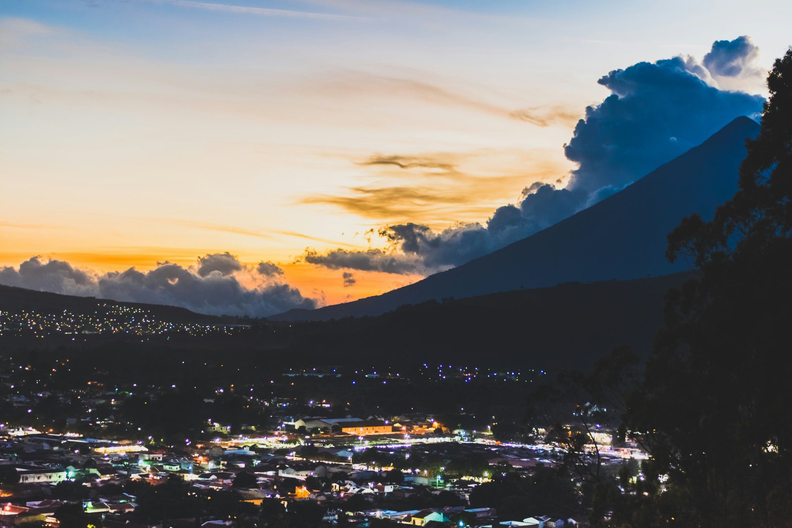 City lights of Antigua Guatemala spread below at dusk with Acatenango and Fuego silhouetted against the orange sky