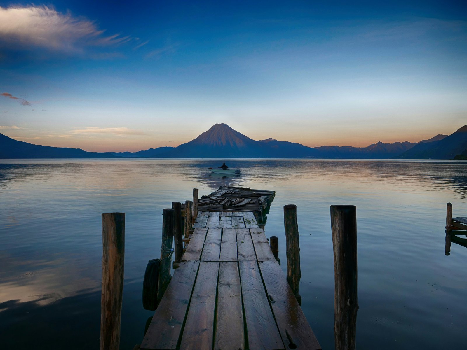 Broken wooden dock stretching into Lago de Atitlan at dawn with a conical volcano reflected in the still blue water