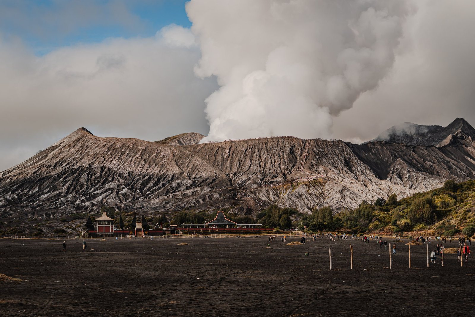 The temple at the foot of Bromo, small against the crater and its steam. Hikers crossing the sand are tiny.