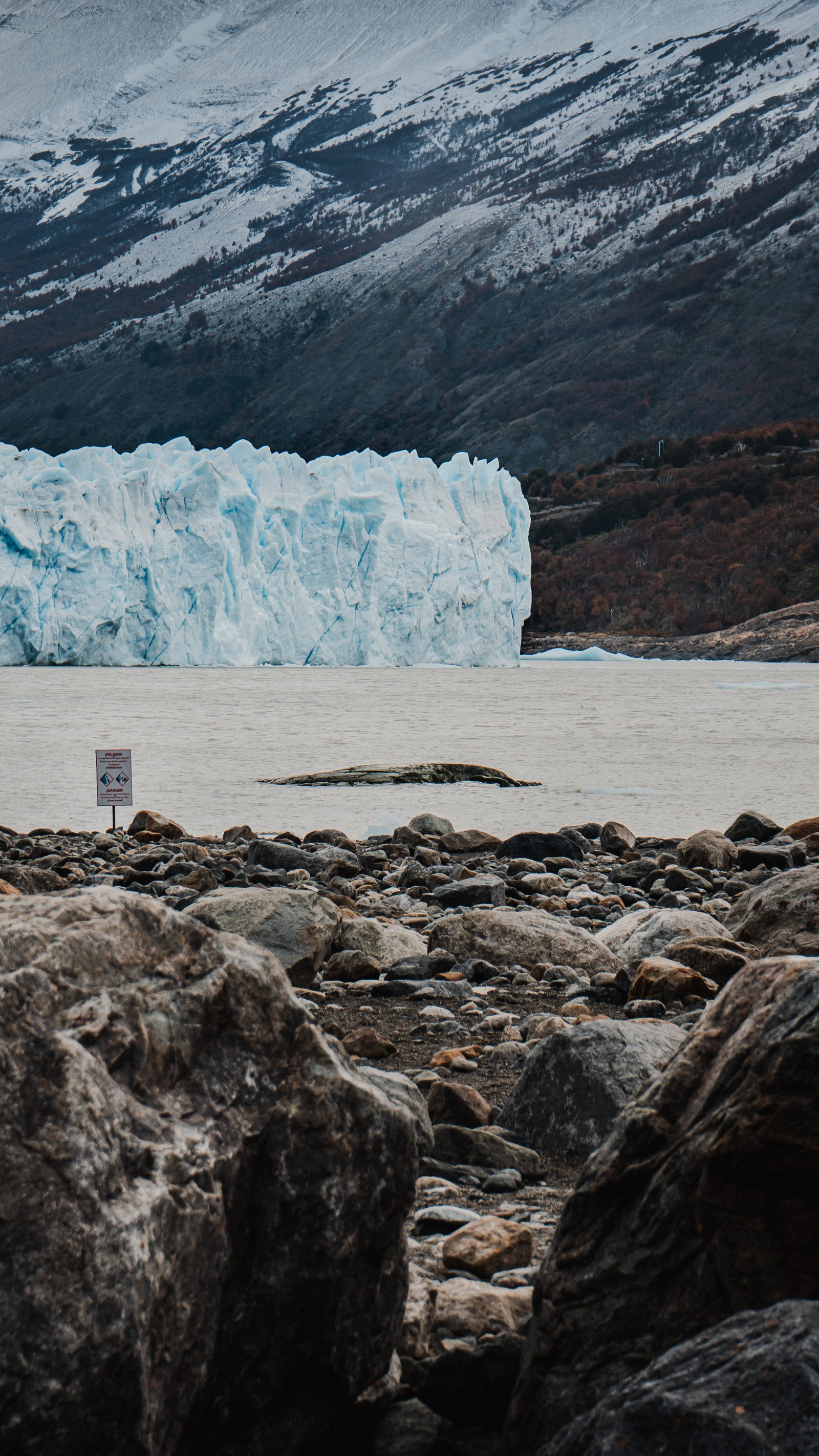 Rocky shore below the glacier. A warning sign at the water's edge.