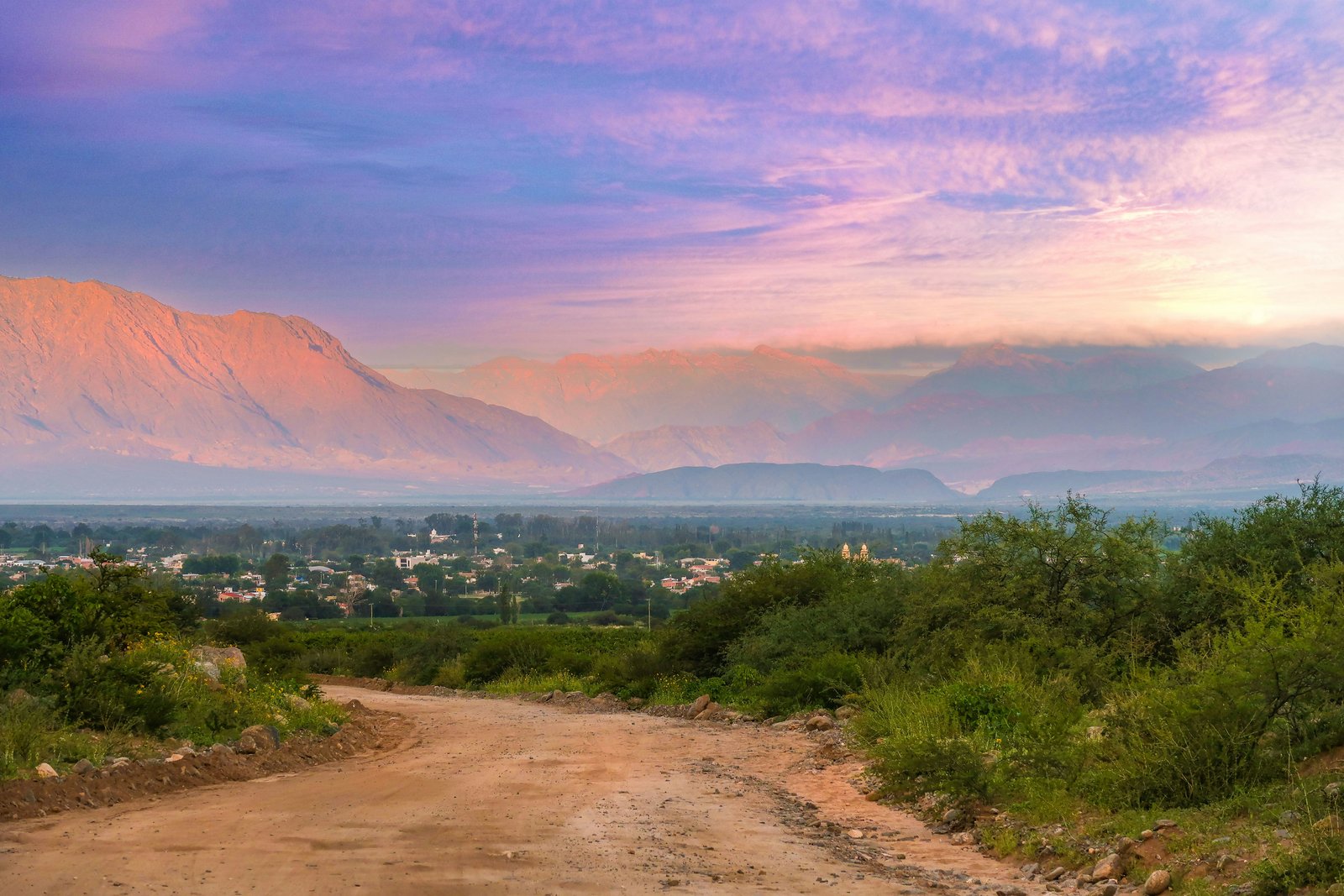 Dirt road descending toward a valley town at dusk, purple and pink Andean sky
